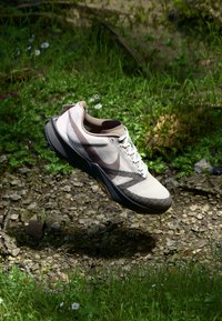 Basket de sport beige clair avec une tige en maille texturée, semelle gris foncé et lacets blancs, présentant un logo subtil en brun foncé sur le côté.