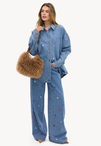 Mujer con camisa azul de mezclilla de gran tamaño y pantalones de pierna ancha decorados con lentejuelas de colores, sosteniendo un bolso grande y esponjoso de color marrón.
