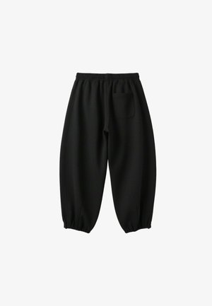Femme portant une veste blanche zippée avec des rayures noires, un crop top noir et un pantalon noir ample sur un fond blanc uni.