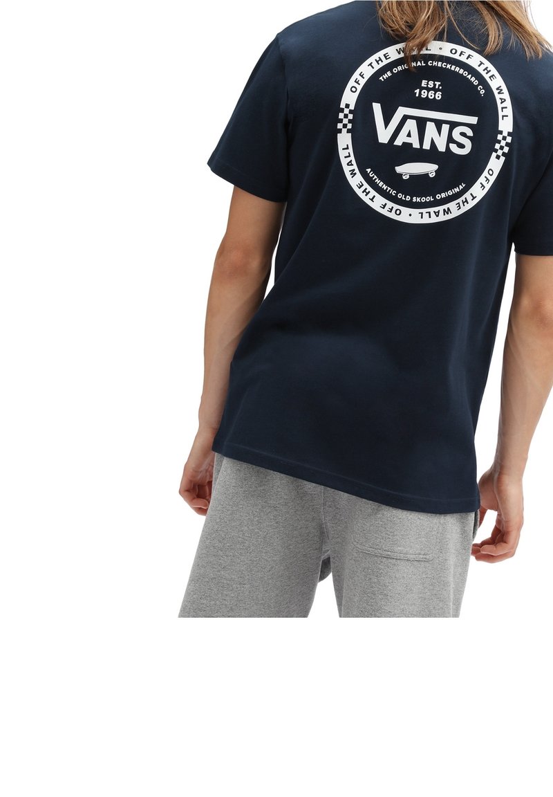 Personne portant un t-shirt Vans bleu marine avec un logo circulaire et un graphisme de skateboard au dos, associé à un pantalon de survêtement gris clair.