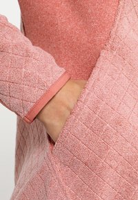 Różowy sweter z pikowanego materiału o teksturowanym wykończeniu i gładkim mankiecie. Materiał jest miękki, z detalem kieszeni po boku.