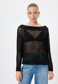 Zwarte gebreide trui met een open maasontwerp en een wijde halslijn, voorzien van lange mouwen en een geribde zoom, gecombineerd met lichtblauwe jeans.