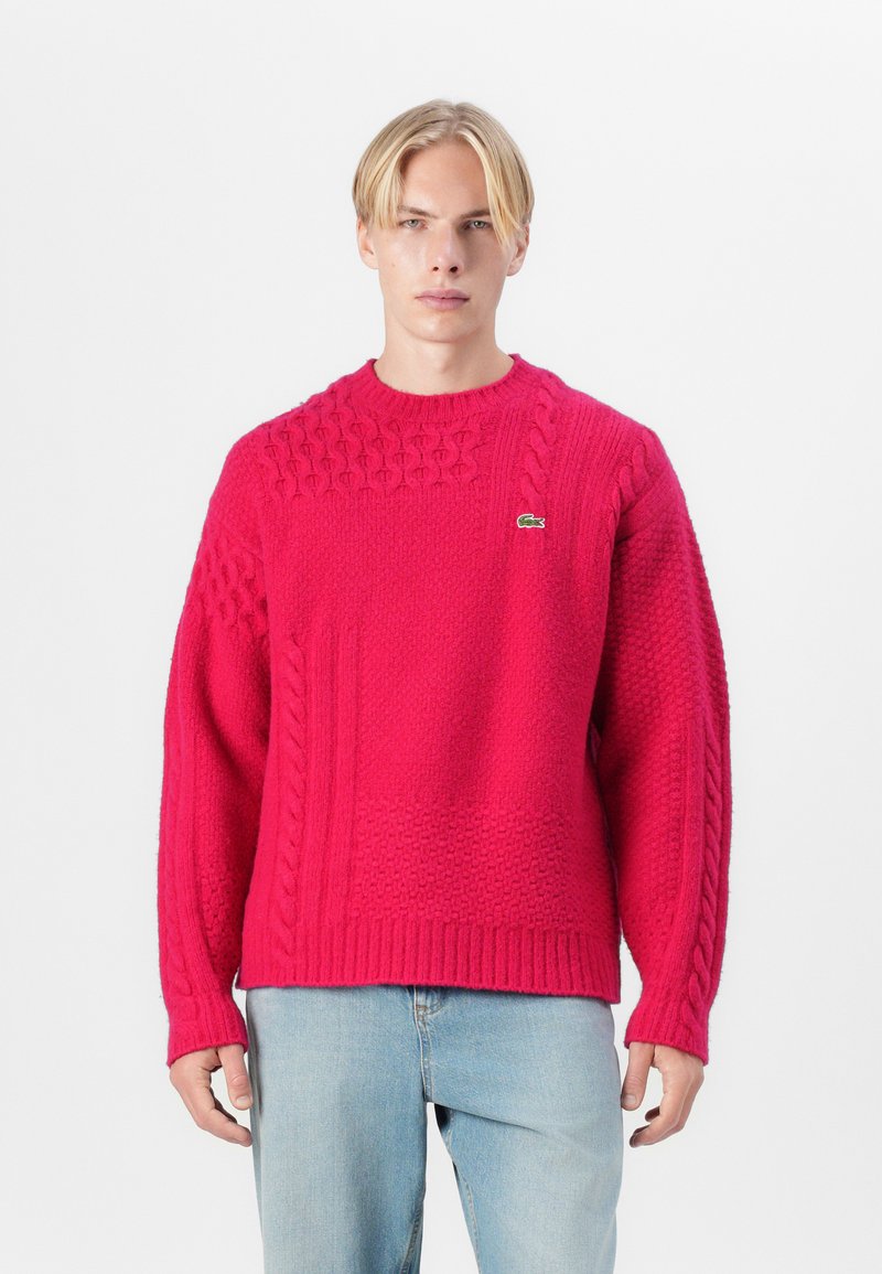 Masīva adījumā rozā džemperis. Ar strukturētām rakstām formām, apaļu izgriezumu un ribotiem piedurknēm un apakšmalu. Lacoste logo uz krūtīm.