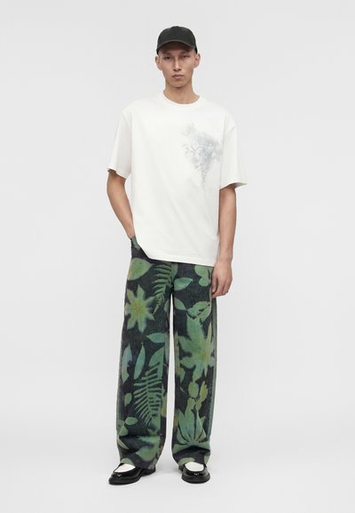 Νέος άνδρας που φοράει μαύρο καπέλο, λευκό κοντομάνικο πουκάμισο με διακριτικό σχέδιο, φαρδιά πράσινα και μαύρα φλοράλ παντελόνια και μαύρα και λευκά παπούτσια.