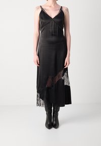 Personne portant une nuisette en satin noir avec bordure en dentelle et des bottes noires pointues sur un fond clair uni.