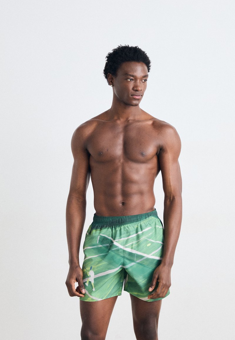 Grønne svømmeshorts med et bladmønster, elastisk talje og et diskret hvidt accentdesign. Materialet ser let og åndbart ud.
