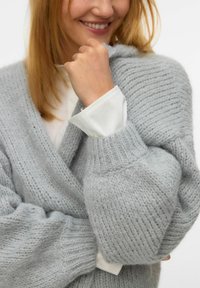 Cárdigan de punto gris claro con escote en V profundo, puños acanalados y textura suave, superpuesto a una blusa blanca con puños visibles.