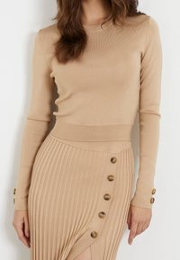 Beige ribbed knit top dengan lengan panjang, menampilkan kerah bulat dan aksen kancing di lengan. Rok pendukungnya berlipat dengan detail kancing.
