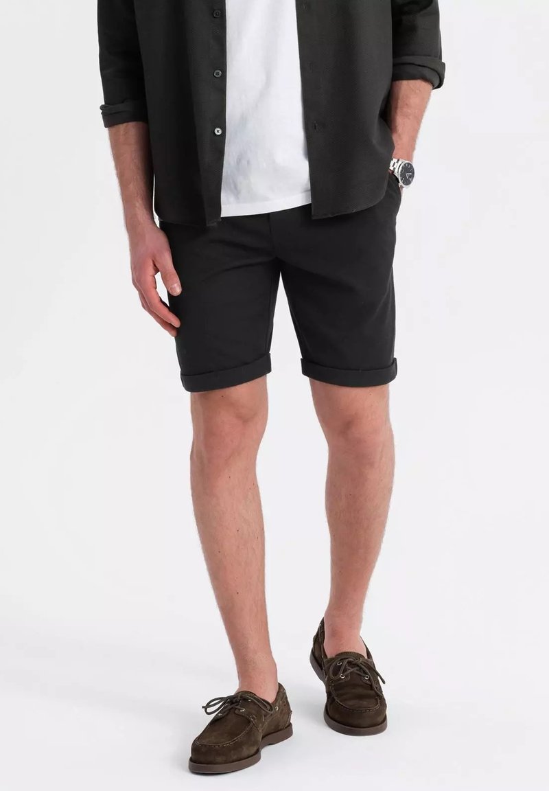 Schwarze Shorts mit umgeschlagenen Säumen, kombiniert mit einem weißen T-Shirt und einem schwarzen Hemd mit Knöpfen. Braune Wildleder-Bootsschuhe barfuß getragen.