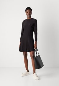 Melnas garās piedurknes kleitas ar nedaudz izplestu apakšmalu, kurai ir neliels logo uz krūtīm, ierīkošana ar melnu somu un baltām čībām.