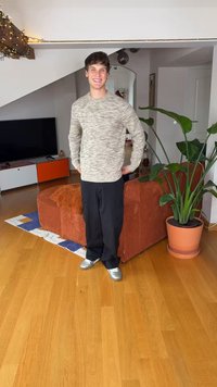 Ljusgrå, texturerad tröja, svarta byxor och silvriga sneakers. Står i ett rum med trägolv, en stor växt och en rund soffa.