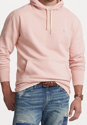 Mand iført en lys pink hættetrøje med forlomme og falmede blå jeans med lapper og et brunt fletbælte.