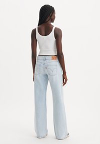 Jean large taille haute bleu clair avec un design classique de poches arrière et un patch en cuir, porté avec un débardeur blanc.