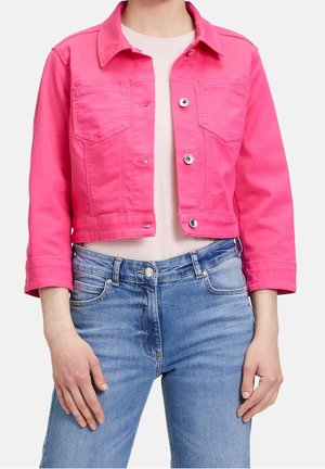 Personne portant une veste courte rose vif avec des boutons argentés sur un haut blanc et un jean bleu, montrée de la poitrine aux hanches.