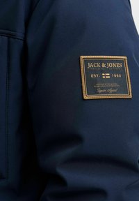 Marineblå jakke med struktureret stof. Har et rektangulært guld- og marineblåt mærke med mærkets navn og etableringsår på skuldren.