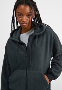 Tmavě šedá fleecová mikina s zipem v přední části, kapucí na šňůrku a předními kapsami. Obsahuje decentní logo na hrudi a texturovanou tkaninu.