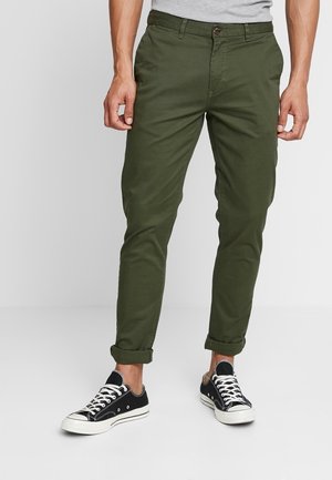 Olivgrüne Chinos mit schmaler Passform, ausgestattet mit einem Knopfverschluss, Fronttaschen und aufgerollten Säumen, kombiniert mit schwarzen Leinenschuhen.