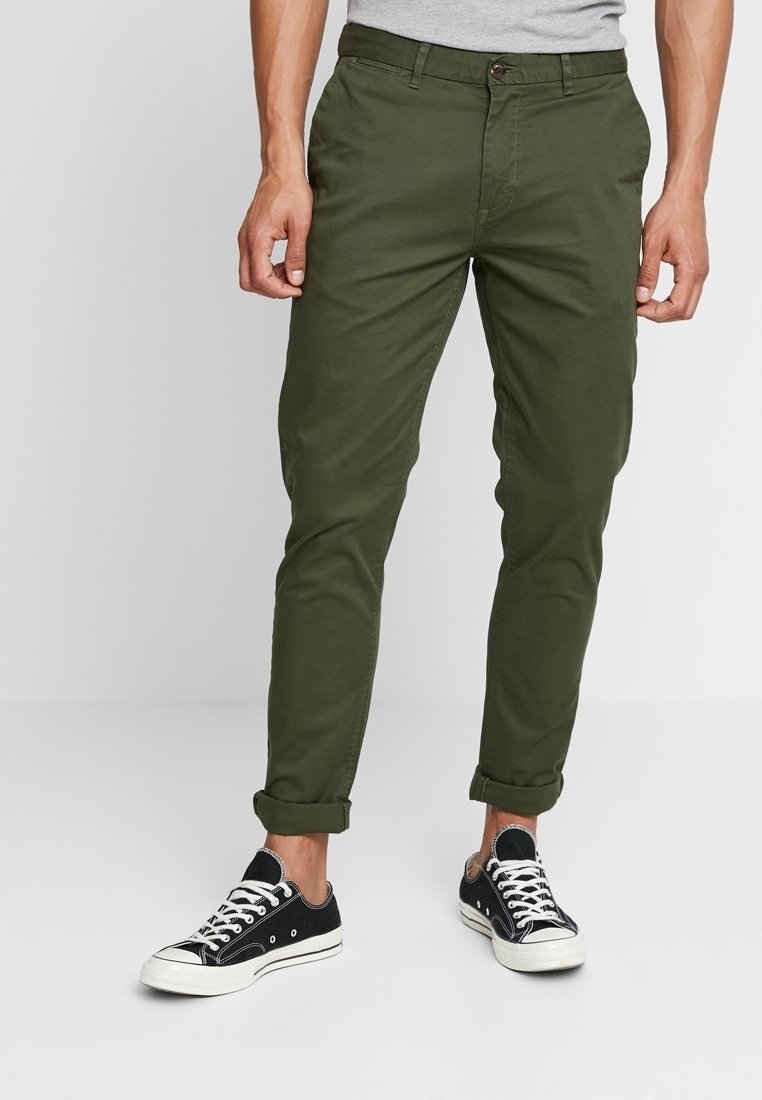 Olivově zelené chinos kalhoty s tapered střihem, se zapínáním na knoflík, předními kapsami a ohrnutými manžetami, spárované s černými plátěnými teniskami.