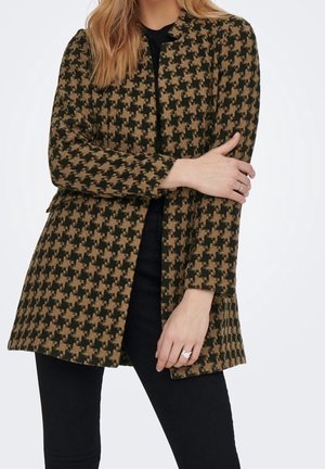 Woman wearing a brown and black houndstooth patterned long coat over black pants, with arms crossed and a ring on her left hand.