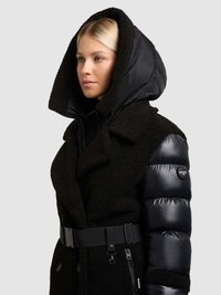 Fekete pufi kabát, bársonyos, texturált külsővel, túméretes kapucnival és övvel a deréknál. Feltűnő cipzáras zsebek és logós díszítések.