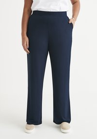 Marineblauwe, wijde broek van soepele stof met een elastische tailleband en zijzakken, gecombineerd met beige instapschoenen.