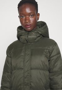 Olivengrøn pufferjakke med høj krave, der har et quiltet design, skinnende tekstur og trykknaplukning foran.