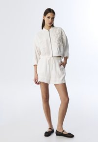Veste courte texturée blanche avec fermeture éclair et poche à rabat, associée à un short blanc avec taille à cordon, et des chaussures plates noires.
