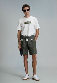 Witte katoenen t-shirt met een print, groene shorts, grijze trui om de taille geknoopt, en witte sneakers met een bruine zool.