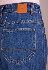Modré džíny z denimu s koženou etiketou s nápisem "CLOSED" na zadním pase. Výrazné oranžové prošívání a zaoblené zadní kapsy.