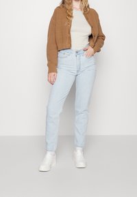 Cardigan cropped a coste marrone sopra una t-shirt a righe chiare, abbinato a jeans a gamba dritta blu chiaro e sneakers bianche.