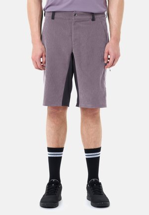 Uomo con pantaloncini viola con pannelli interni neri, scarpe da ginnastica nere e calzini neri con due strisce bianche, in piedi contro uno sfondo bianco.