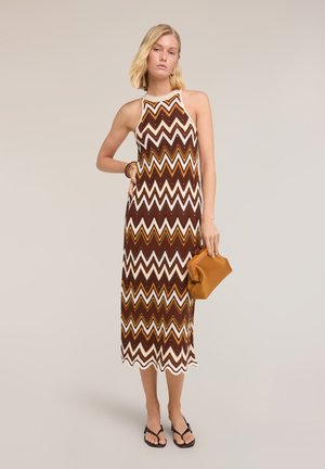 Femme portant une robe sans manches à motif zigzag marron, blanc et jaune, tenant une pochette marron et portant des sandales noires.