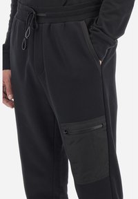 Zwarte joggingbroek van zachte stof met een elastische tailleband, voorzien van een ritszak op de rechterdij in een contrasterend matte materiaal.