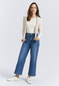 Széles szárú, közép-kék farmernadrág, magas derekú, elülső zsebekkel, krémszínű hosszú ujjú felsővel és fehér tornacipővel párosítva.