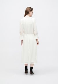 Dlouhé bílé šaty s projmutým pasem, průhlednými rukávy a volánkovým límcem; mají rovný střih a délku po kolena. Viditelné černé průhledné ponožky a podpatky.