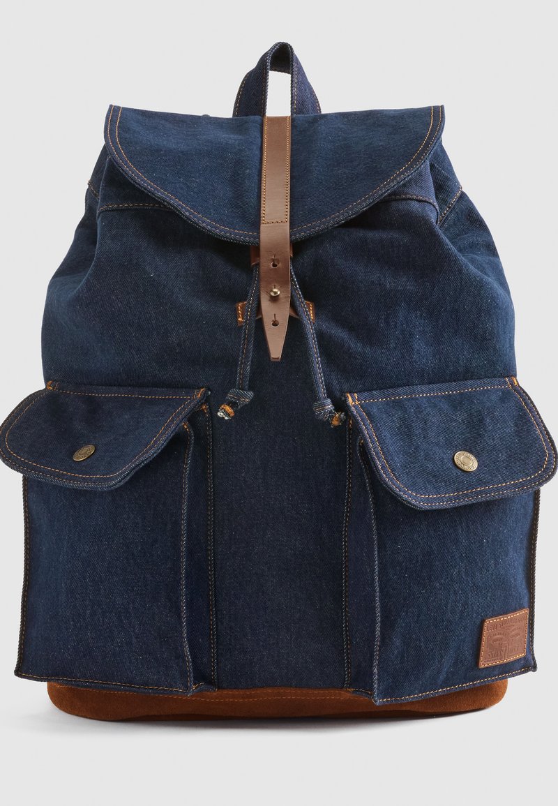 Zaino in denim di colore blu scuro, con dettagli in pelle, chiusura a patta, due tasche frontali e una base marrone. Presenta dettagli di cucitura a contrasto.