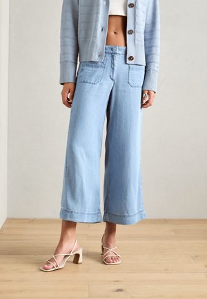 Kvinna iklädd ljusblå vida beskurna jeans, en ljusblå kashmirtröja, vit crop top och beige strap-on blockklacksandaler stående på trägolv.