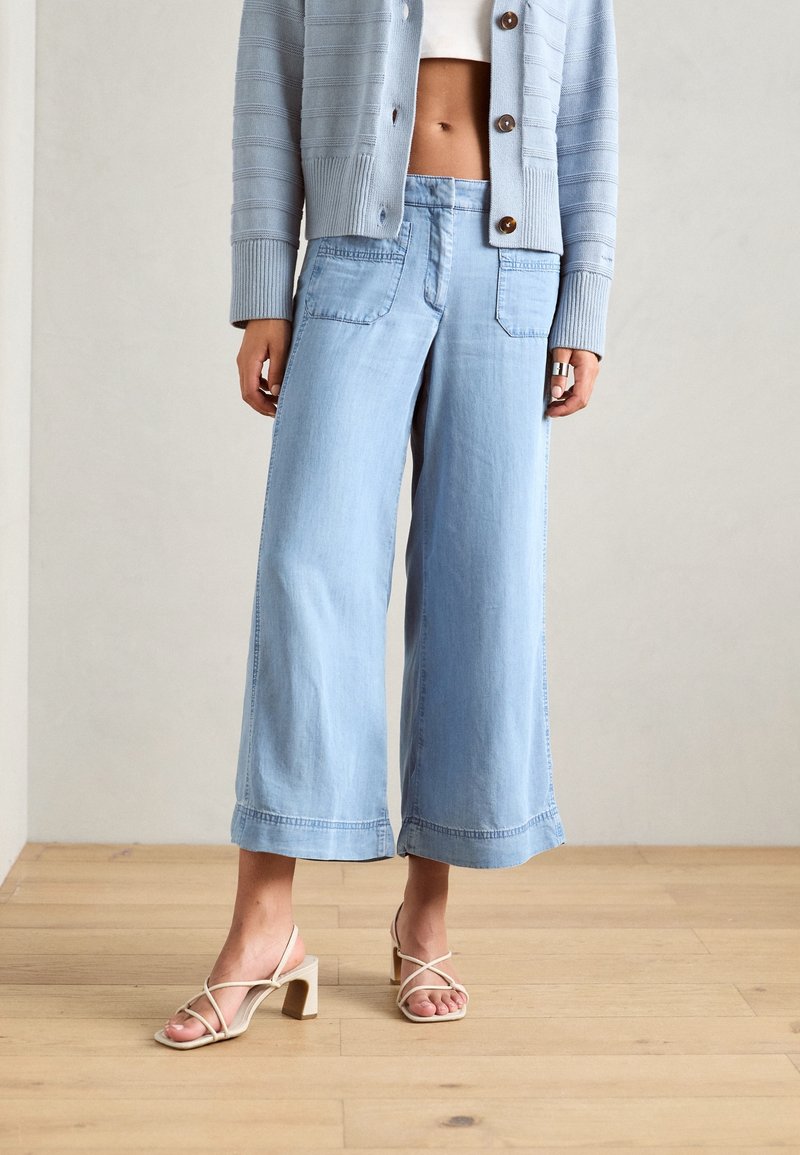 Žena oblečená v světle modrých širokých cropped džínech, světle modrém kardiganu, bílém crop topu a béžových sandálech na blokovém podpatku stojí na dřevěné podlaze.