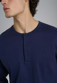 Uomo che indossa una maglietta henley blu navy con tre bottoni scuri, mostrato dalle spalle al mento su uno sfondo grigio uniforme.