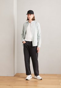 Camicia button-up verde chiaro sopra una maglietta bianca, abbinata a jeans scuri e larghi e sneakers bianche con dettagli neri. Cappellino nero indossato.