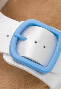 Witte nep leren sandaalband met een blauwe plastic gesp, voorzien van twee gaten voor aanpassing en een gestructureerd oppervlak. Zandkleurige voetbed zichtbaar.