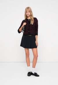 Leopard patterned burgundy cardigan with black trim, paired with a black pleated mini skirt. Black loafers and white crew socks complete the look.