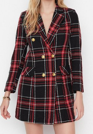 Kobieta ubrana w czerwoną i czarną kratowaną sukienkę-marynarkę z podwójnym rzędem złotych guzików i klapkowymi kieszeniami, stojąca na białym tle.