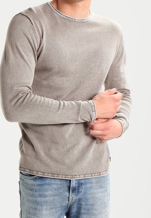 Chemise grise à manches longues en tissu doux, avec un col rond et des détails de couture visibles, associée à un jeans en denim bleu clair.