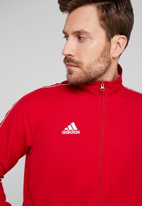 Roter Reißverschlussjacke aus glattem Stoff, mit weißen Streifenakzenten an den Schultern und einem weißen Adidas-Logo auf der Brust.