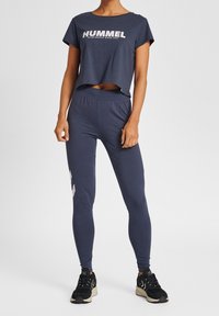Marinblå crop top med "HUMMEL"-logotyp, kombinerad med åtsittande marinblå leggings med ljusgrå chevrondetaljer och svarta träningsskor.