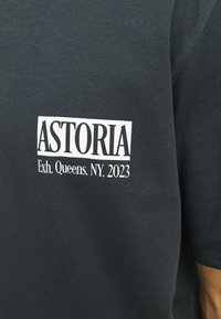 Tamsiai pilka medvilninė marškinėliai su baltu spaudiniu "ASTORIA" ir "Exh. Queens, NY. 2023" šiuolaikiniu, ryškiu šriftu.