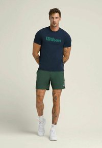 Hombre con tatuajes que lleva una camiseta navy de Wilson, pantalones cortos verdes, calcetines blancos y zapatillas blancas, de pie contra un fondo neutro.