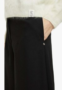 Pantaloni neri con una tasca laterale dotata di una cerniera argento. La parte superiore è un maglione leggero e soffice con un'etichetta che recita "Qui dentro fa caldo."