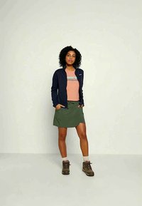 Sieviete stāv ar rokām kabatās, ģērbta tumši zilā jakā, persiku krāsas Patagonia T-kreklā, zaļā svārkā, baltās zeķēs un brūnās pārgājēju zābakos.